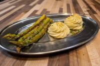 Two piped Potato Kisses Bramborové Pusinky swirls beside roasted asparagus on a metal tray