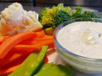 Glass bowl of creamy herbed Ranch Dip surrounded by carrot sticks, broccoli, cauliflower, and snap peas.