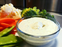 Glass bowl of Ranch Dip surrounded by cauliflower, broccoli, red pepper, and snap peas.