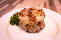 Rustic Crushed Potatoes with Bacon and Onion Mound of Rustic Crushed Potatoes with Bacon and Onion, topped with crispy bacon and browned onion, on a white plate.