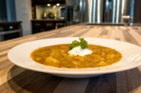 A bowl of Czech Sauerkraut Soup Zelňačka with potato chunks, a dollop of sour cream, and a parsley garnish, set on a striped wooden countertop.