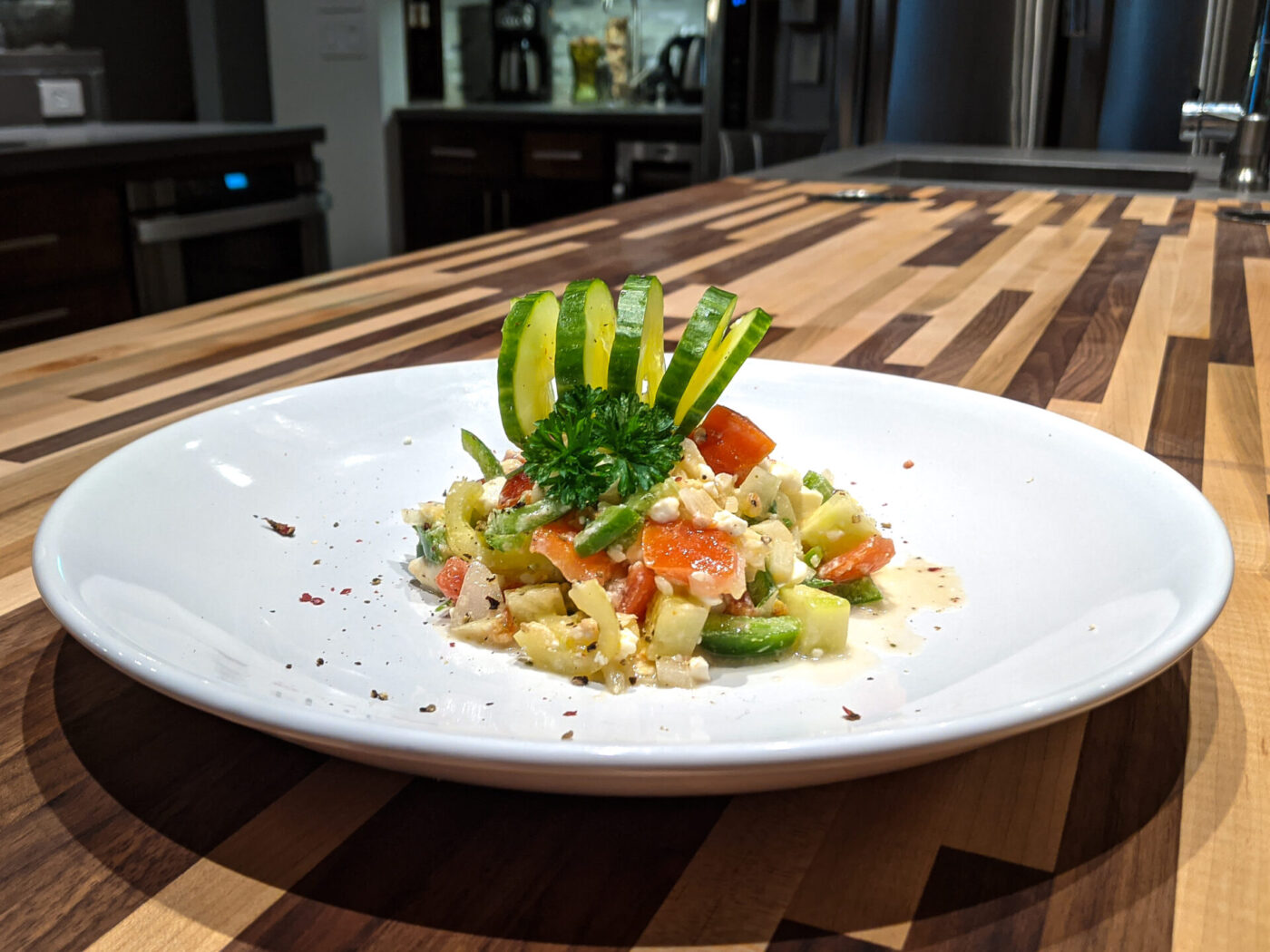 Shopska Salad Diced Shopska Salad on a white plate, topped with a cucumber ribbon fan and herb sprig, on a striped wood counter.