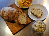 Glass bowl of Artichoke Spinach Dip beside a slice of bread spread with it, plus sourdough loaf and tortilla chips.