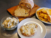 Artichoke Spinach Dip in a glass bowl, spread on bread, with sliced bread and tortilla chips.