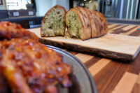 Sliced Twice-baked Herb Bread with green herb flecks on a wooden board beside glazed meat