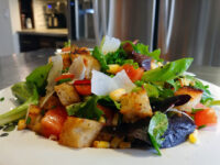 Vinaigrette Salad with mixed greens, tomatoes, corn, croutons, and shaved cheese on a white plate.