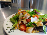 Vinaigrette Salad with mixed greens, tomatoes, corn, croutons, and shaved cheese on a white plate.