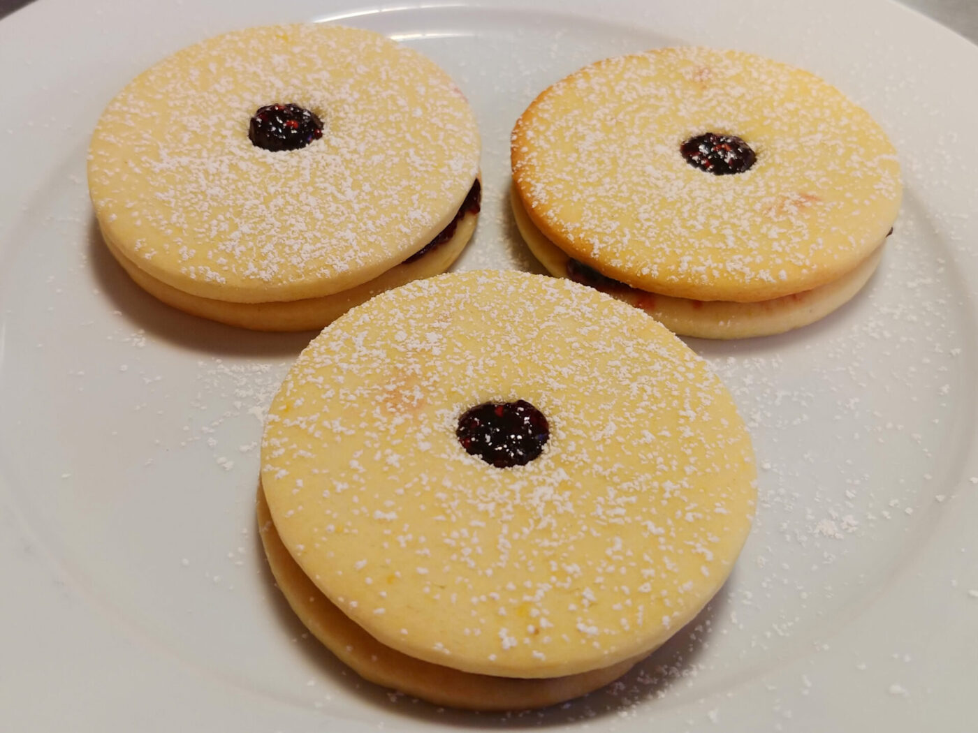 Three Linzer Tarts Cookies (Linecké Cukroví) dusted with powdered sugar, dark jam visible at centers, on a white plate.