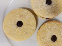 Three powdered-sugar-dusted Linzer Tarts Cookies (Linecké Cukroví) with dark fruit filling on a white plate.
