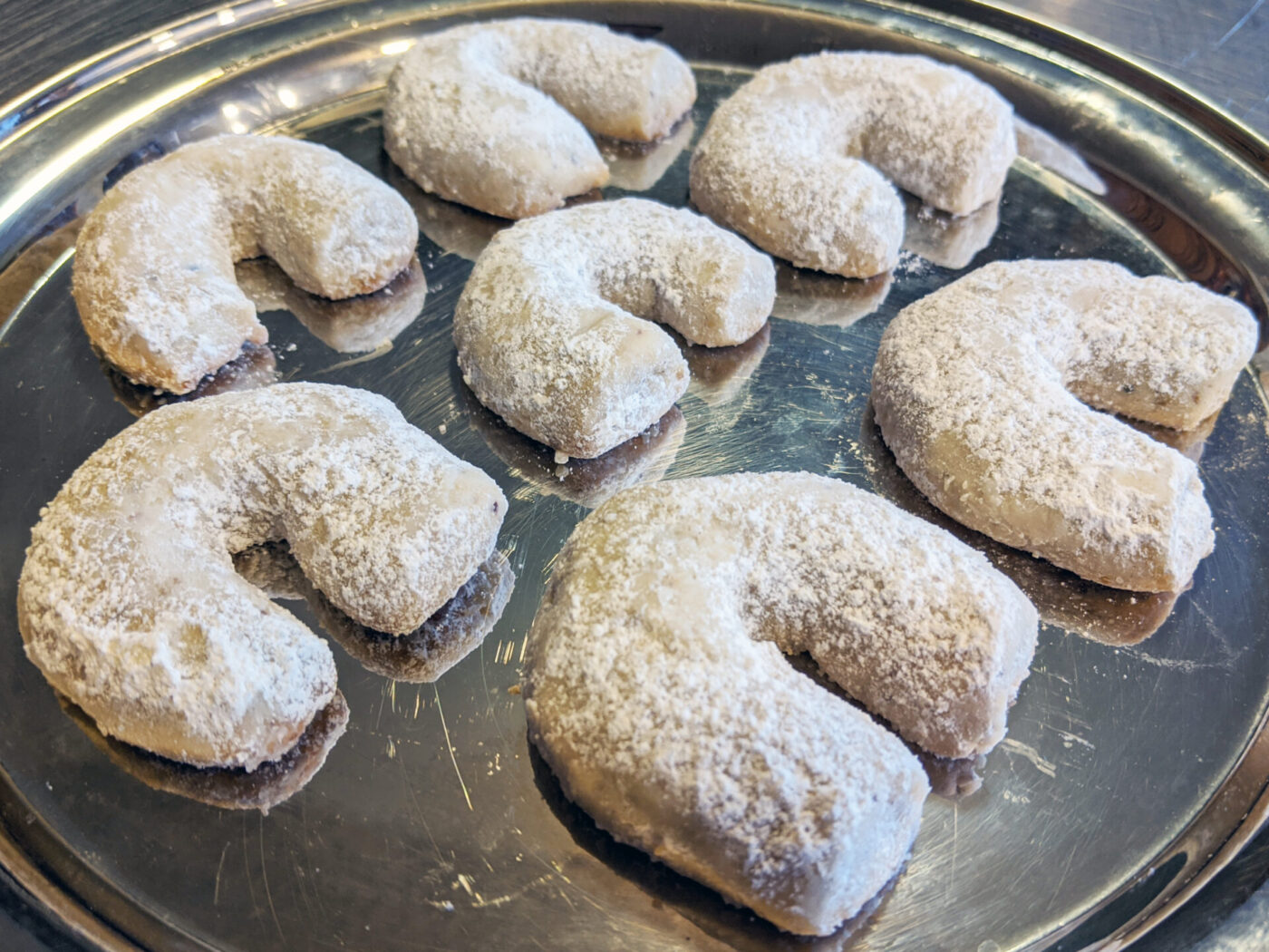 Seven Vanilla-walnut Crescents (vanilkové rohlíčky) dusted with powdered sugar on a round metal tray.