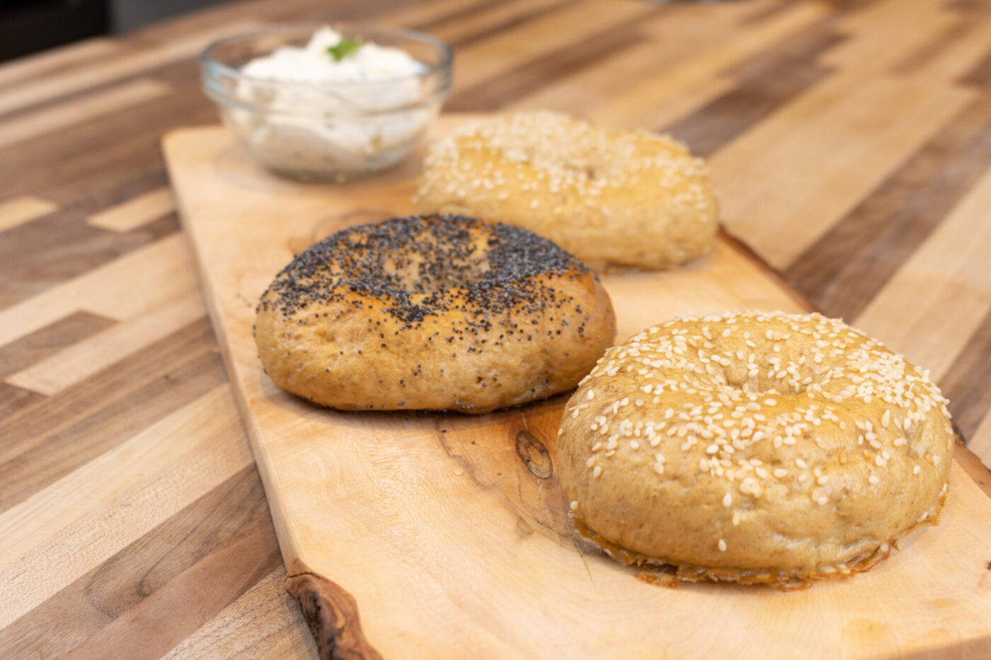 Three seeded Bagels, one topped with poppy seeds, two with sesame, on a wooden board beside a cream cheese dip.