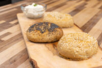 Three seeded Bagels, one topped with poppy seeds, two with sesame, on a wooden board beside a cream cheese dip.