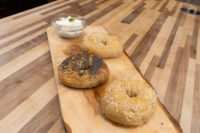 Three bagels, two sesame-topped and one poppy seed-topped, on a wood board beside a bowl of cream cheese