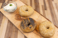 Three bagels, two with sesame seeds and one with poppy seeds, on a wooden board beside a bowl of cream cheese
