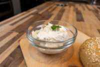 Glass bowl of Creamy Spread with Garlic and Chives topped with a herb leaf, beside a sesame seed roll.