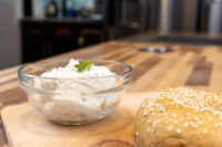 Glass bowl of Creamy Spread with Garlic and Chives, garnished with herb, beside a sesame seed roll