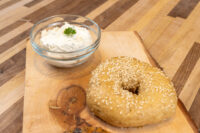 Sesame seed bagel on a wooden board beside a glass bowl of cream cheese with parsley.
