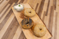 Three seed-topped bagels on a wooden board next to a small bowl of cream cheese dip.