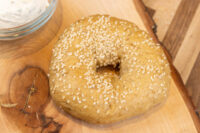 A sesame seed-covered bagel on a wood board, with a bowl of herbed cream cheese nearby