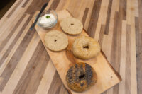 Wooden board with four sliced bagel halves, plain, sesame, and poppy seed, beside cream cheese.