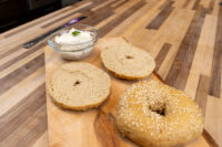 Small bowl of Creamy Spread with Garlic and Chives, herb-flecked, beside bagel halves on a wooden board