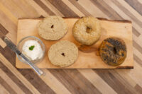 Four halved bagels, plain, sesame, and poppy seed, on a wood board beside a bowl of herbed cream cheese