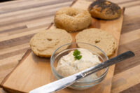 Glass bowl of Creamy Spread with Garlic and Chives, herb-topped, beside halved bagels and a knife on a wooden board.