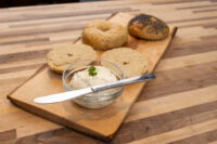 Assorted bagel halves on a wooden board next to a small bowl of Creamy Spread topped with a sprig of herb.