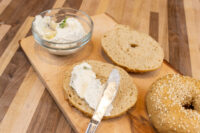 Halved bagel spread with cream cheese beside a whole sesame bagel and a bowl of cream cheese on a wooden board.