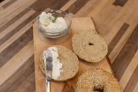 Sliced Bagel halves spread with cream cheese beside a sesame Bagel on a wooden board.