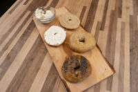 Four halved bagels with seeds and cream cheese on a wooden board beside a bowl of herbed cream cheese.