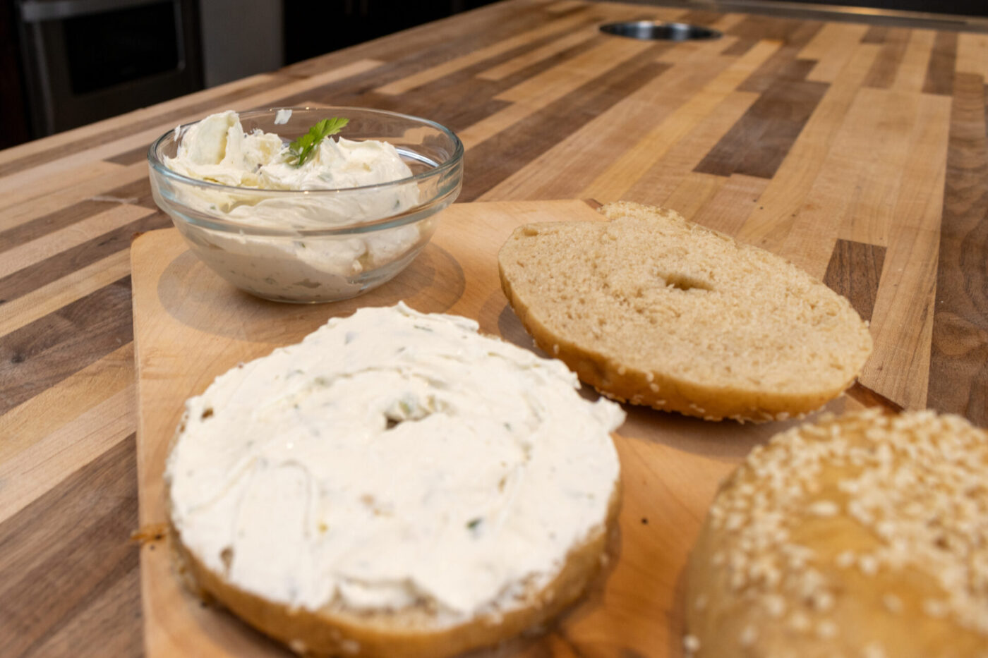 Bagel halves on a wooden board, one spread with herbed cream cheese, beside a bowl of the same spread.