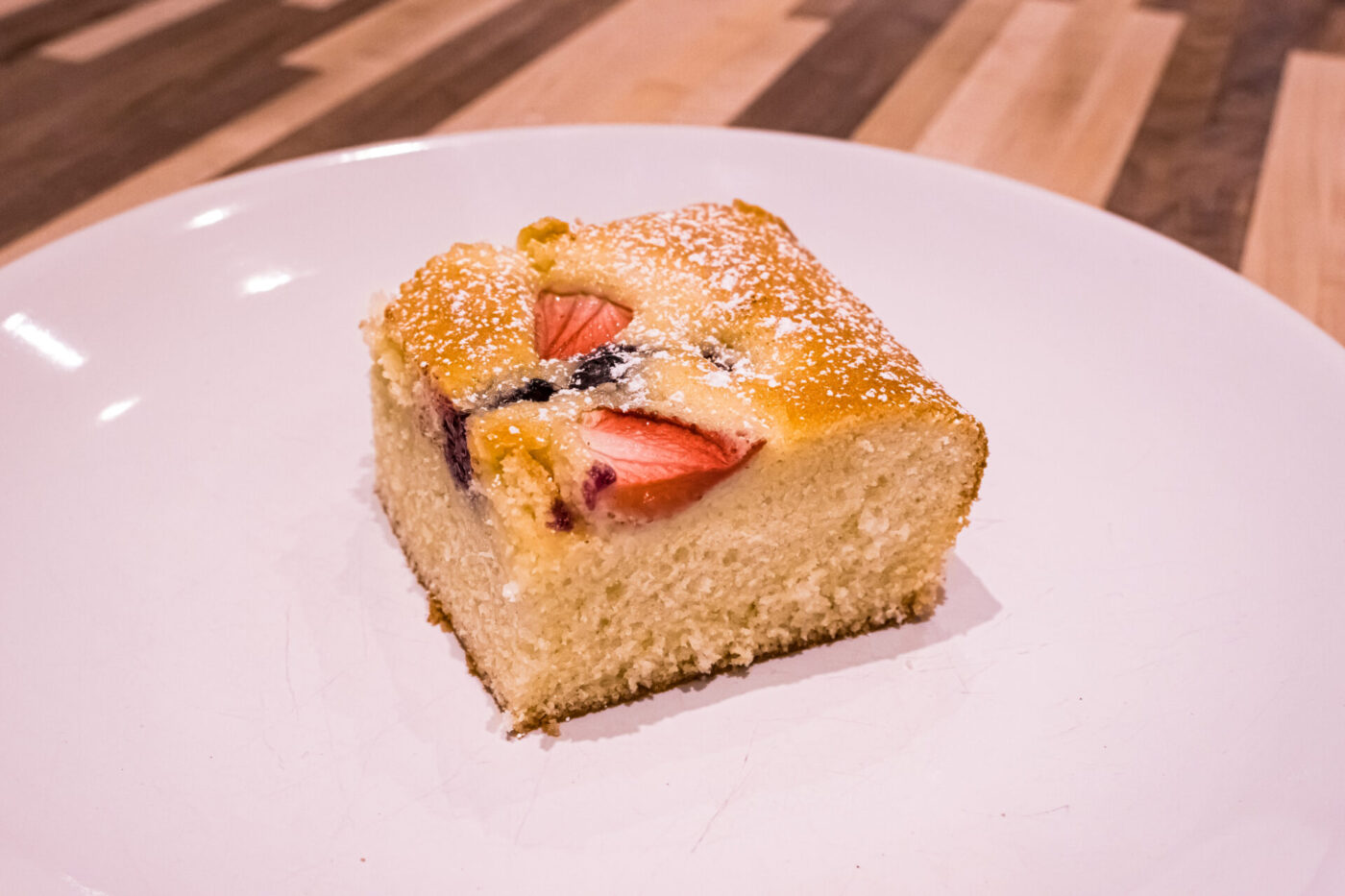 Square slice of Bublanina (Czech Bubble Cake) with strawberries, dark berries, and powdered sugar on a white plate.