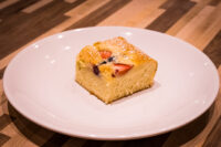 Square slice of Bublanina (Czech Bubble Cake) with strawberry and berry pieces, dusted with powdered sugar, on a white plate.