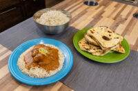 Butter Chicken with rice on a blue plate, beside a bowl of rice and a green plate of flatbread.