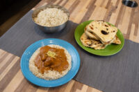 Butter Chicken sauce with meat pieces over rice on a blue plate, beside rice and naan bread.