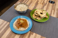 Butter Chicken with lemon zest over rice on a blue plate, beside a rice bowl and stacked flatbreads.