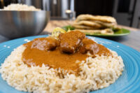 Butter Chicken pieces in thick orange sauce over herbed rice on a blue plate, naan visible behind.