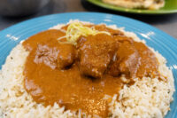 Blue plate of Butter Chicken: saucy meat chunks in orange-brown gravy over herbed rice, topped with scallions.