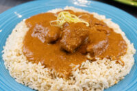 Butter Chicken chunks in orange-brown sauce over herbed rice on a blue plate, topped with lemon zest strips.