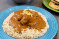 Butter Chicken pieces in thick tomato sauce over herbed rice, garnished with citrus zest, on a teal plate.