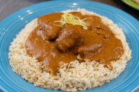 Butter Chicken pieces in reddish sauce topped with zest, served over herbed rice on a blue plate