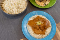 Butter Chicken with pale garnish on rice, blue plate, beside rice pot and naan on green plate.
