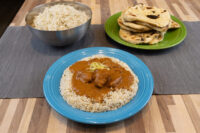 Butter Chicken with meat in brown curry sauce over rice on a blue plate, beside rice and naan.