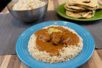 Butter Chicken over rice on a blue plate, with a bowl of rice and flatbreads beside it.