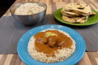 Blue plate of Butter Chicken over rice, with a metal bowl of rice and flatbreads nearby.