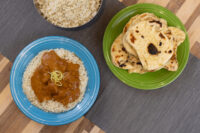 Butter Chicken curry with rice on a blue plate beside naan bread and a bowl of rice.