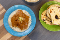 Butter Chicken with rice and citrus zest on a blue plate, served beside folded naan on a green plate.