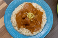 Blue plate of Butter Chicken, tomato sauce and chicken pieces over herbed rice, topped with citrus zest.
