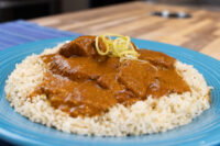 Butter Chicken with tender meat chunks in tomato sauce over rice, garnished with lemon zest, on a blue plate.
